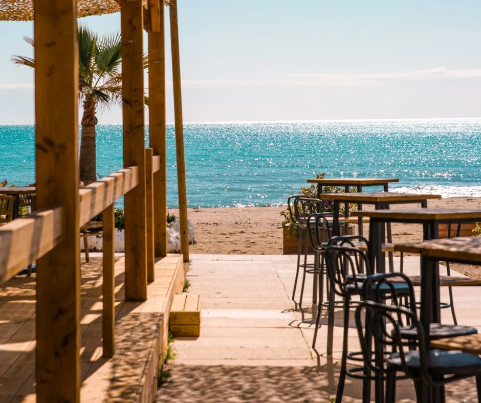 A sunlit Costa del Sol beach with a traditional wooden chiringuito grilling espetos