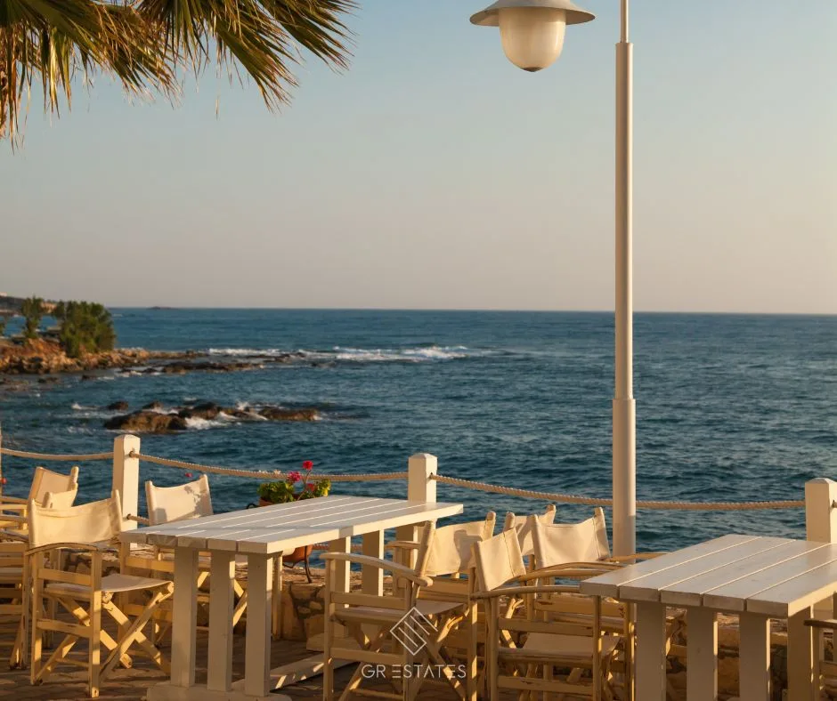 Terrazza di un caffè sul lungomare all'ora dorata, con tavoli di legno bianchi e sedie da regista dietro una ringhiera di corda, una foglia di palma, un lampione, gerani rossi e un mare blu e calmo che s'infrange sugli scogli